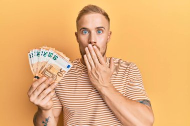 Elinde 50 avroluk banknotlar tutan genç beyaz bir adam ağzı eliyle kapatıyor, şaşırmış ve hata yapmaktan korkuyor. şaşırmış ifade 