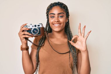 Saç örgülü, elinde antika fotoğraf makinesi olan genç bir Afro-Amerikan kadını parmaklarıyla işaret veriyor, gülümseyerek mükemmel bir sembol oluşturuyor. 