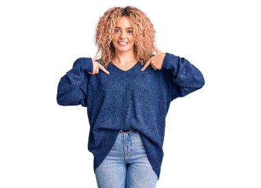 Kıvırcık saçlı, sıradan bir kazak giyen, kendine güvenen, gülümseyen, kendini gururlu ve mutlu parmaklarıyla gösteren genç sarışın kadın.. 