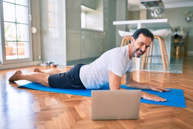 Orta yaşlı, sakallı ve esnek bir adam evde egzersiz yapıyor bilgisayardaki spor videolarına bakıyor.