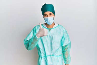 Cerrah üniforması giyen ve tıbbi maske takan genç yakışıklı adam elleriyle mutlu bir jest yapıyor. Kameraya bakıp başarıyı gösteren ifade. 