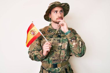 Kafkasyalı genç adam kamuflaj üniforması giyiyor İspanyol bayrağı taşıyor ciddi bir yüz ifadesi var eli çenesinde soru sormayı düşünüyor, kafa karıştırıcı bir fikir. 