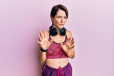 Kısa saçlı, spor kıyafetleri ve kulaklıkları olan genç esmer kadın tiksindirici tepki verdiği için tiksindirici ve tiksindirici bir ifade takınıyor. Eller havada. 