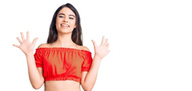 Esmer genç kız gündelik giysiler giyer ve parmaklarını on numara ile gösterir ve gösterirken kendinden emin ve mutlu bir şekilde gülümser.. 