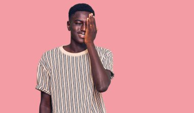 Genç Afro-Amerikan adam günlük kıyafetler giyiyor. Bir gözünü eliyle kapatıyor. Yüzünde kendine güvenen bir gülümseme ve sürpriz bir duygu var.. 