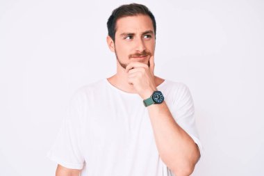 Elleri çenesinde gündelik beyaz tişört giyen genç ve yakışıklı bir adam soru sormayı düşünüyor, düşünceli bir ifade. Düşünceli bir yüzle gülümsüyor. şüphe kavramı. 