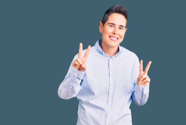 Kısa saçlı, iş kıyafetleri giymiş, gülümseyen, parmaklarını zafer işareti yaparken gösteren genç bir kadın. İki numara.. 