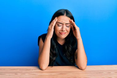 Güzel esmer, gündelik kıyafetler giyen genç bir kadın elleri başının üstünde masada oturuyor, stres yüzünden baş ağrısı çekiyor. migren ağrısı. 
