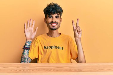 Üzerinde mutluluk mesajı olan İspanyol tişörtü giyen genç adam masada oturmuş yedinci parmaklarını gösterirken kendinden emin ve mutlu bir şekilde gülümsüyor.. 