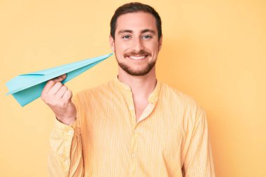 Elinde kağıt uçak tutan yakışıklı genç adam pozitif ve mutlu görünüyor. Dişlerini gösteren kendine güvenen bir gülümsemeyle ayakta duruyor ve gülümsüyor. 