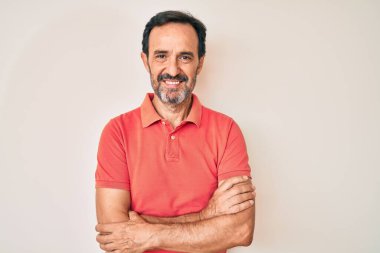 Orta yaşlı İspanyol bir adam sıradan kıyafetler giyip kollarını kavuşturup kameraya bakarken gülümsüyor. pozitif kişi. 