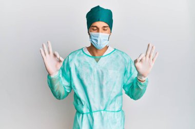 Cerrah üniforması ve tıbbi maske takan genç yakışıklı adam rahatla ve gülümse gözleri kapalı parmaklarıyla meditasyon hareketi yaparken. yoga konsepti. 