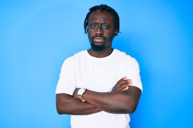 Örgülü, sıradan beyaz tişörtlü, kuşkucu ve gergin, yüzünde çapraz kollu tasvip etmeyen bir ifade olan Afro-Amerikalı genç bir adam. negatif kişi. 