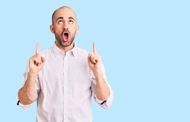 Zarif gömlek giyen genç yakışıklı adam şaşırmış ve şaşırmış bir şekilde yukarı bakıyor ve parmaklarıyla işaret ediyor ve kollarını kaldırıyor.. 
