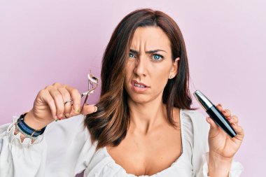 Young brunette woman holding eyelash curler clueless and confused expression. doubt concept.  clipart