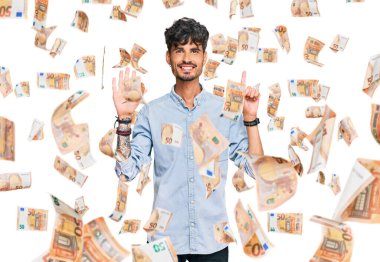 Latin asıllı genç adam gündelik giysiler giyip altı numaralı parmaklarıyla işaret ederken kendinden emin ve mutlu bir şekilde gülümsüyor..