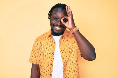 Saçları örgülü, el hareketleriyle gülümseyen, mutlu yüzüyle parmaklarının arasından bakan Afro-Amerikan genç adam.. 