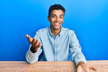 Genç, yakışıklı, Afro-Amerikan bir adam, gündelik giysiler içinde masada oturmuş neşeli bir şekilde gülümsüyor, dostça karşılıyor, olumlu ve kendinden emin selamlıyor. 
