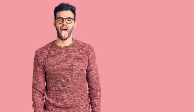 Kış süveteri giyen ve gözlük takan genç, yakışıklı İspanyol adam komik bir ifadeyle dilini dışarı çıkartıyor. duygu kavramı. 