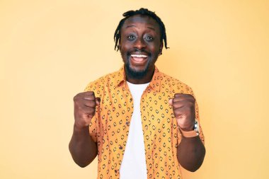 Saçları günlük kıyafetlerle örülmüş genç Afro-Amerikalı adam kollarını kaldırarak ve açık gözlerle başarıyı kutluyor ve şaşırıyor. Kazanan konsept. 