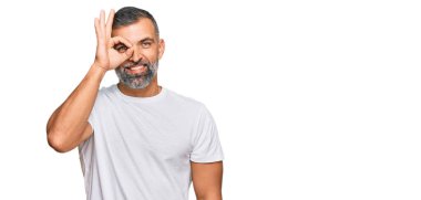 Orta yaşlı, beyaz tişört giyen yakışıklı bir adam el hareketleriyle gülümsüyor ve mutlu yüzüyle parmaklarının arasından bakıyor.. 