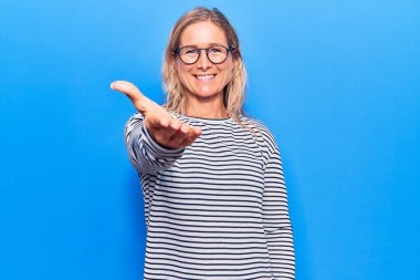 Orta yaşlı, beyaz, çizgili süveter ve gözlük takan sarışın bir kadın dostça bir selamlama ve hoş geldin hediyesi olarak gülümsüyor. başarılı iş. 