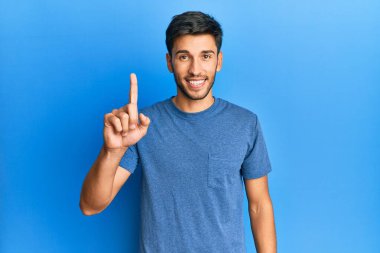 Mavi arka planda sıradan tişörtler giyen genç yakışıklı bir adam bir fikir veya soruyla gülümsüyor ve mutlu bir yüzle işaret ediyor. 