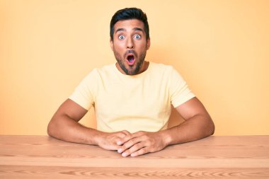 Gündelik giysiler giyen genç İspanyol adam masada korkmuş ve ağzı açık bir şekilde şaşırmış bir şekilde oturuyor. 