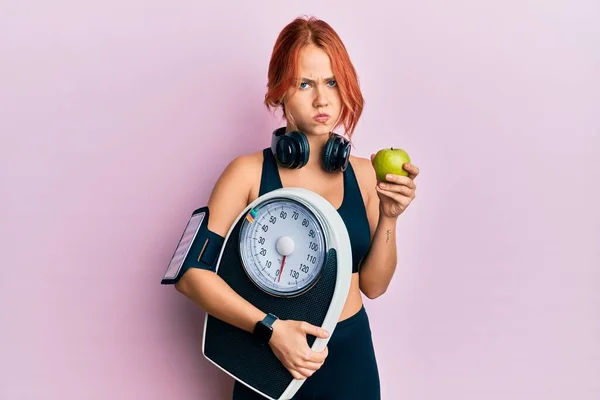 Young beautiful redhead woman holding weight machine to balance weight loss skeptic and nervous, frowning upset because of problem. negative person.