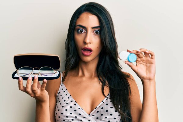 Красивая латиноамериканка, держащая очки и контактные линзы в шокирующем лице, выглядящая скептически и саркастично, удивленная открытым ртом 