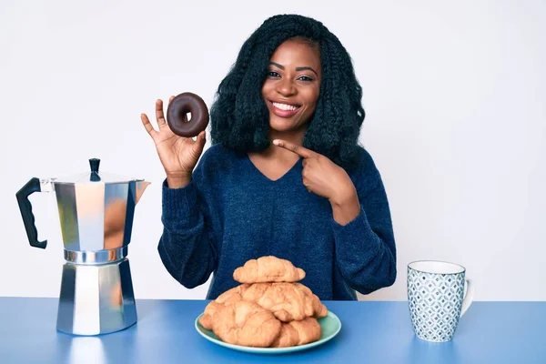 Beautiful african woman eating breakfast holding cholate donut smiling happy pointing with hand and finger