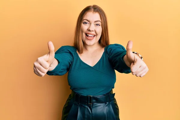 Young beautiful redhead woman wearing elegant and sexy look approving doing positive gesture with hand, thumbs up smiling and happy for success. winner gesture.
