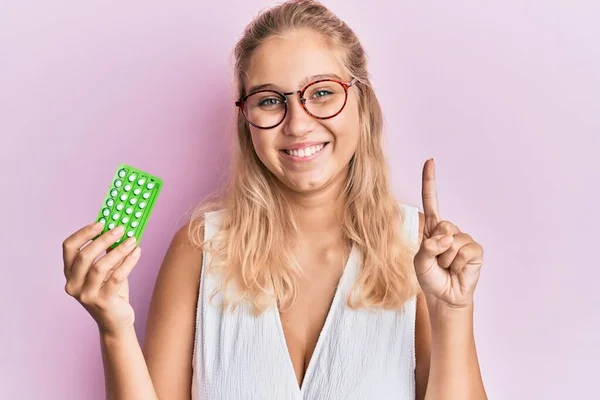 Young blonde girl holding birth control pills smiling with an idea or question pointing finger with happy face, number one