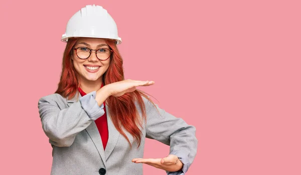 大小記号を示す手で建築家のハードハットジェスチャーを身に着けている若い赤毛の女性は シンボルを測定します カメラを見て笑ってる 概念を測定し — ストック写真