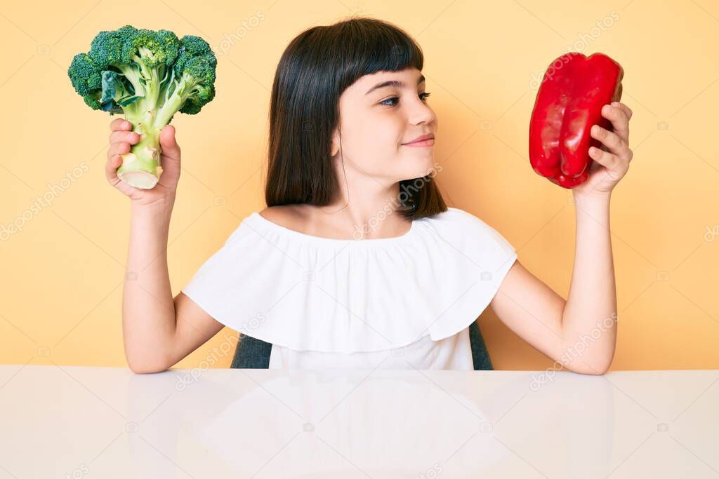 Young little girl with bang holding broccoli and red pepper smiling looking to the side and staring away thinking. 