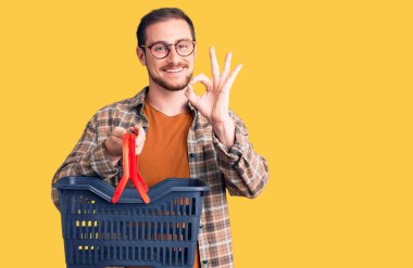 Elinde süpermarket sepeti olan yakışıklı beyaz bir adam parmaklarıyla imza atıyor, gülümseyerek mükemmel bir sembol oluşturuyor. 