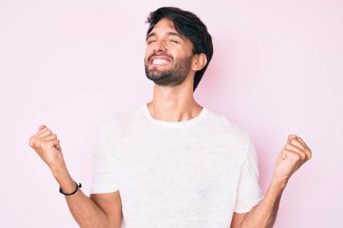 Yakışıklı, beyaz tişört giyen çok mutlu ve heyecanlı kolları havada, gülümseyen ve başarı için bağıran yakışıklı bir İspanyol. kutlama konsepti. 