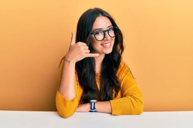 Güzel, esmer, gözlüklü ve günlük giysiler giyen genç bir kadın masaya oturmuş gülümsüyor telefonla el ve parmaklarıyla telefonla konuşuyor gibi hareket ediyor. iletişim kavramları. 