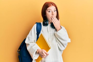 Genç kızıl saçlı kız öğrenci çantası takıyor ve elinde kitapla ağzını kapatıyor. Şok olmuş ve hata yapmaktan korkuyor. şaşırmış ifade 