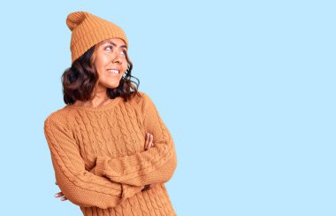 Genç, güzel, melez, yün kazak ve kış şapkası giyen, kolları çapraz ve kendinden emin bir şekilde yana bakan bir kadın. 