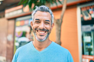 Orta yaşlı, kır saçlı, gülümseyen bir adam şehrin caddesinde mutlu bir şekilde yürüyor..