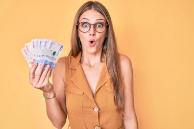 Genç sarışın kız elinde Kolombiya pezosu tutuyor. Korkmuş ve şaşırmış bir şekilde ağzı açık bekliyor. 