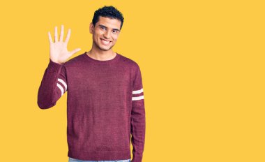 İspanyol görünümlü, sıradan kıyafetler giyen genç adam beş numarayı gösterip işaret ederken kendinden emin ve mutlu bir şekilde gülümsüyor.. 