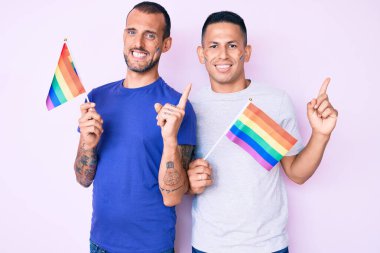 Gökkuşağı lgbtq bayraklarını yan yana tutan iki genç gay el ve parmak ile mutlu bir şekilde gülümsüyor. 