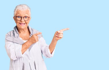 Mavi gözlü, gri saçlı, sıradan giyinmiş, gözlüklü, gülümseyen ve kameraya bakan, iki eliyle ve parmaklarıyla yüzünü gösteren son sınıf güzeli kadın.. 