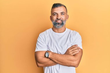 Orta yaşlı, beyaz tişört giyen, kollarını kavuşturup kameraya gülümseyen yakışıklı bir adam. pozitif kişi. 