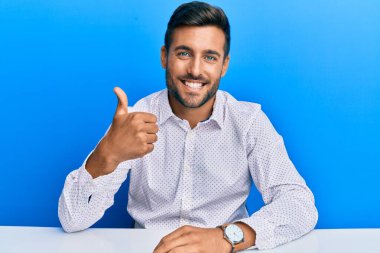 İş kıyafetleri giymiş yakışıklı İspanyol adam masaya oturup el hareketleriyle mutlu mesut el sallıyor. Kameraya bakıp başarıyı gösteren ifade. 