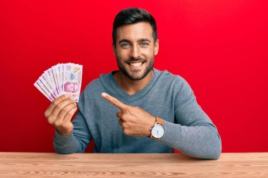 Meksika pezosu tutan yakışıklı İspanyol adam mutlu bir şekilde el ve parmak işareti yapıyor. 