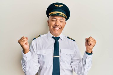 Yakışıklı, olgun, uçak pilotu üniforması giyen mutlu ve heyecanlı bir adam. Kolları havada, gülümseyen ve başarı için bağıran, kazanan bir jest yapıyor. kutlama konsepti. 