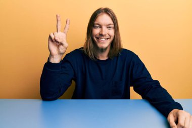 Uzun saçlı, yakışıklı beyaz bir adam masadaki gündelik kıyafetlerini giymiş iki numaralı parmaklarıyla oturmuş kendinden emin ve mutlu bir şekilde gülümsüyor.. 